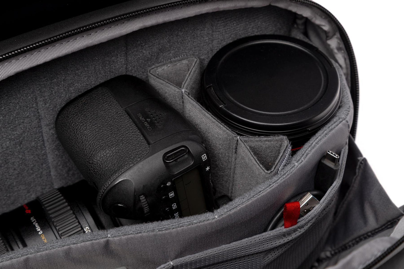 Close-up of a gray camera bag interior with a DSLR camera and lens secured in a padded compartment, alongside a lens cap and small accessories.