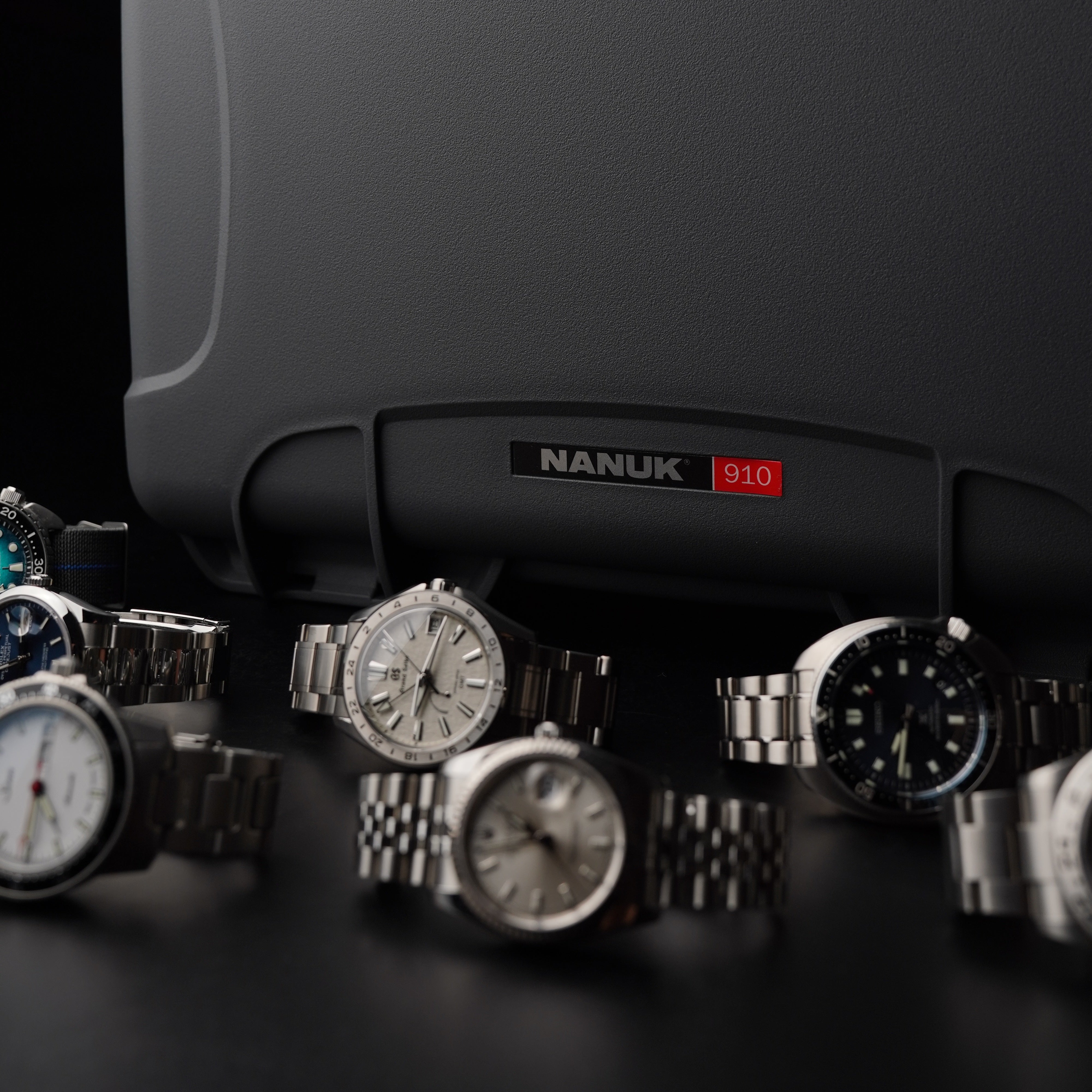 Gray hard-shell carrying case with 'Nanuk' branding, surrounded by various watches on a dark background.