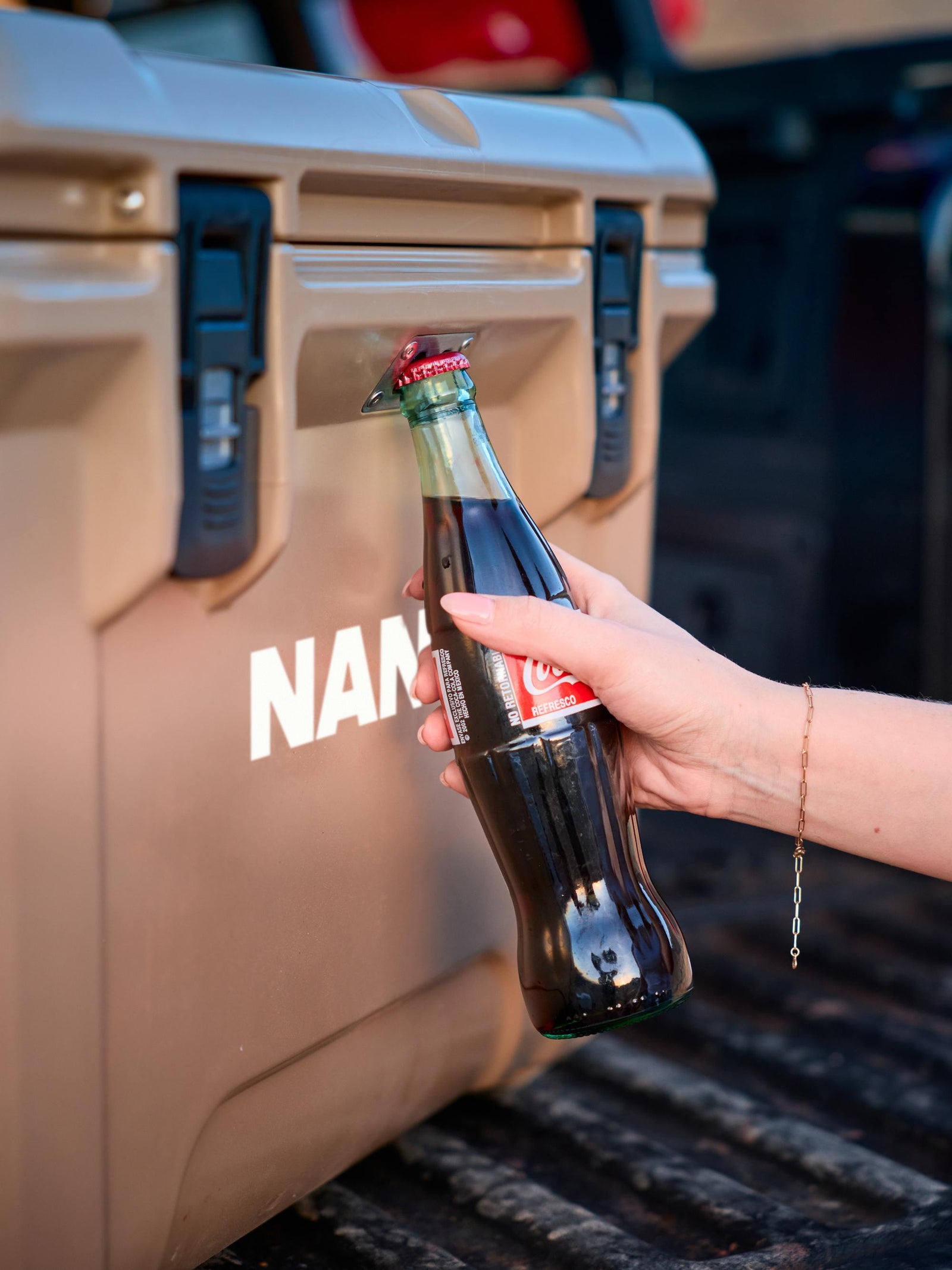 Person opening a bottle of Coca-Cola next to a beige cooler with 'NAN' branding.