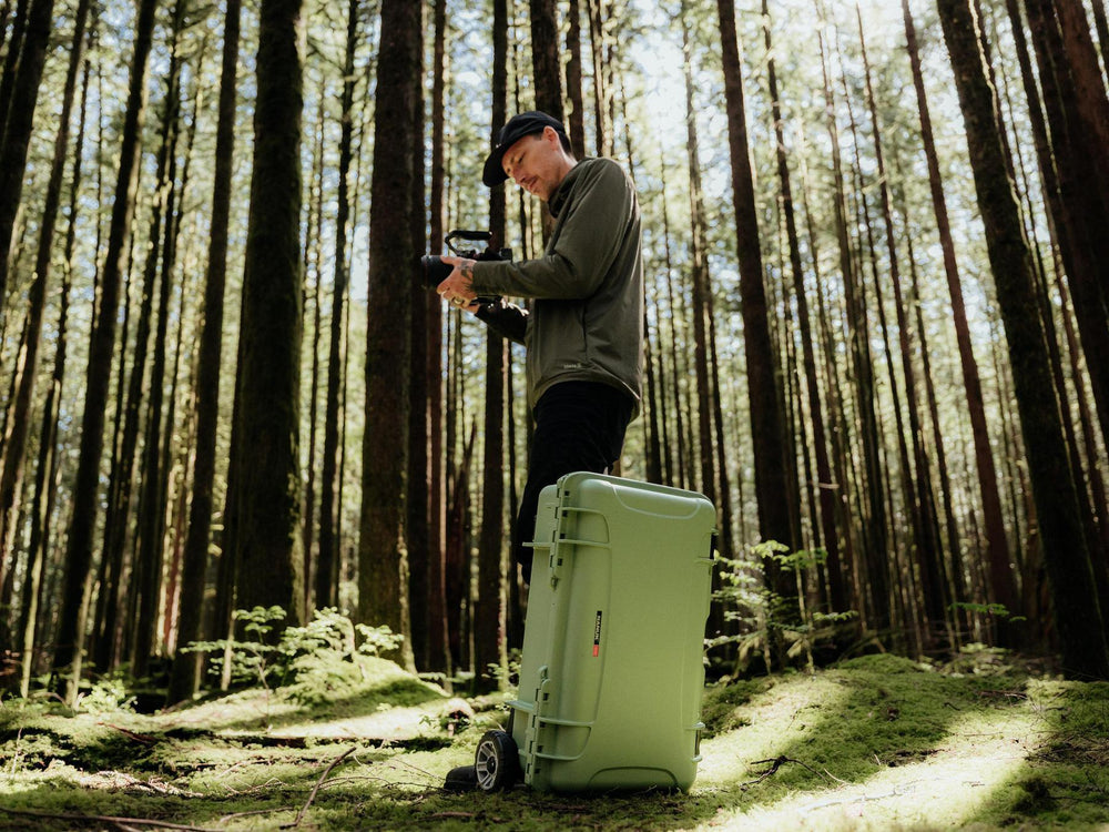 Person setting up a camera in a forest with a large green suitcase.