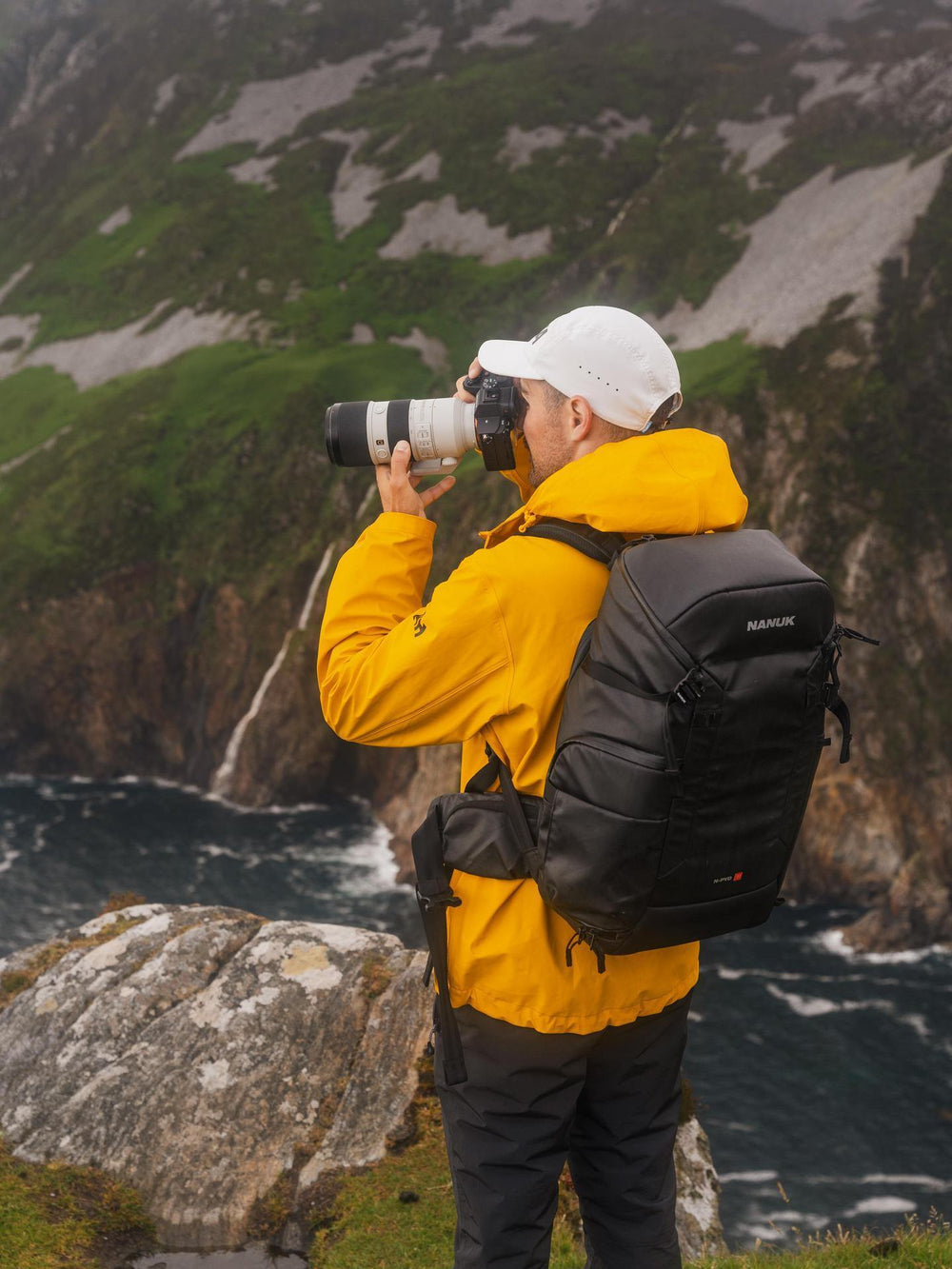 Person in yellow jacket and black backpack taking a photo of a scenic landscape with mountains and water.