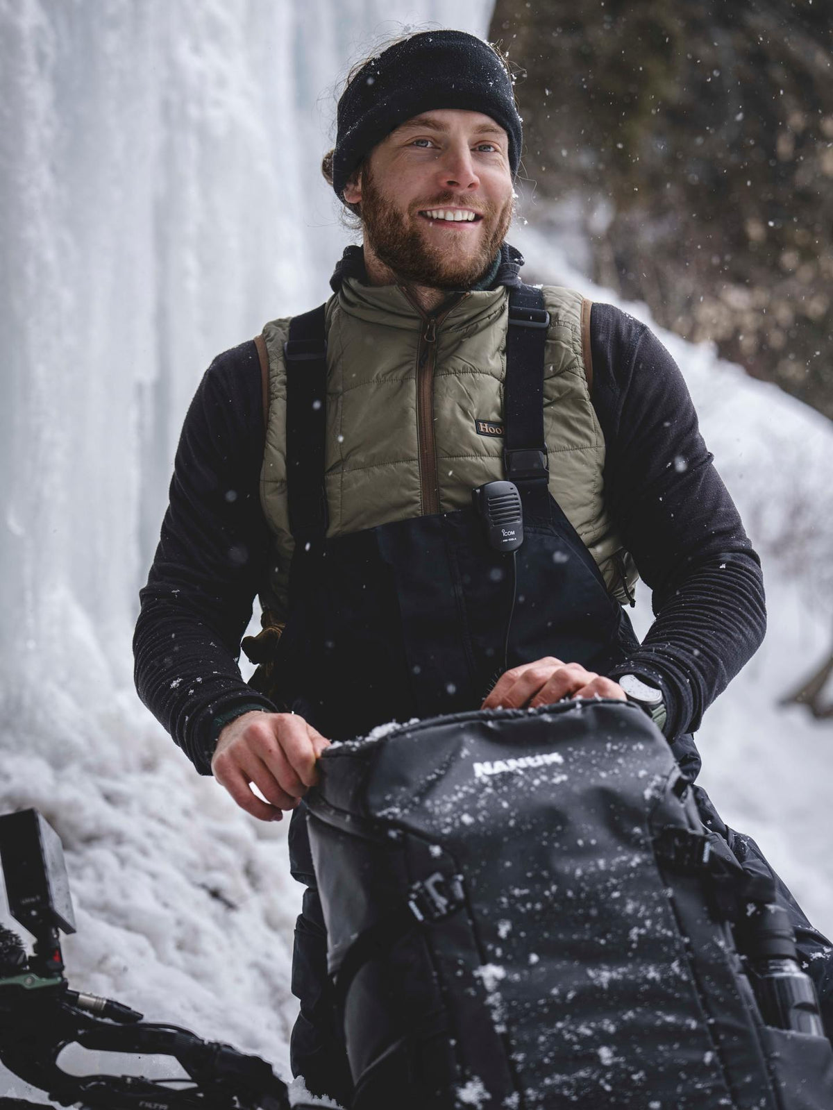 Man in winter clothing with a backpack standing in front of a snowy waterfall.