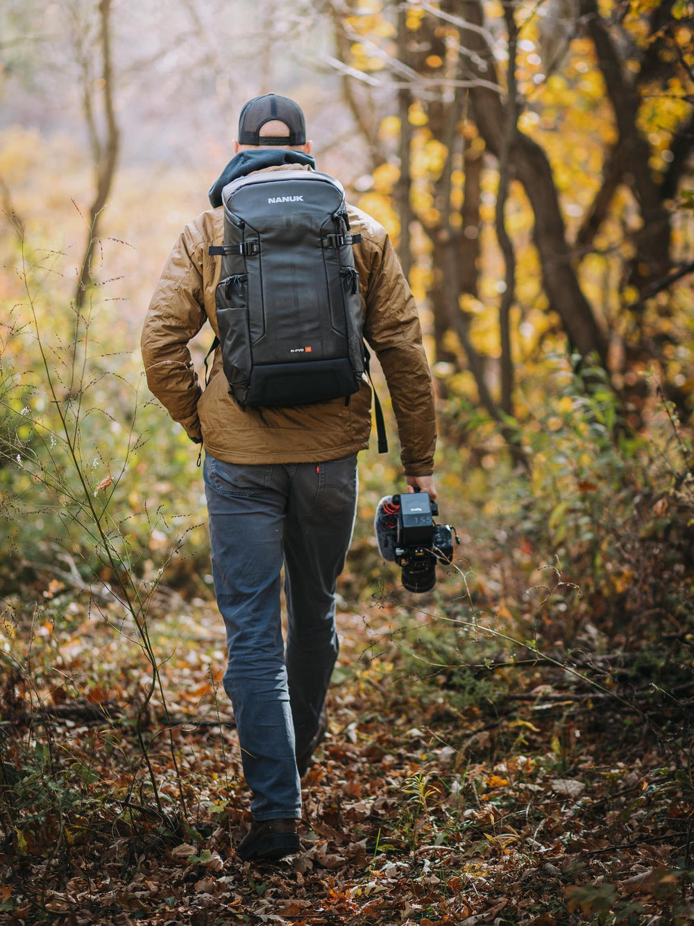 Person walking in a forest with a backpack and camera, wearing a jacket and cap.