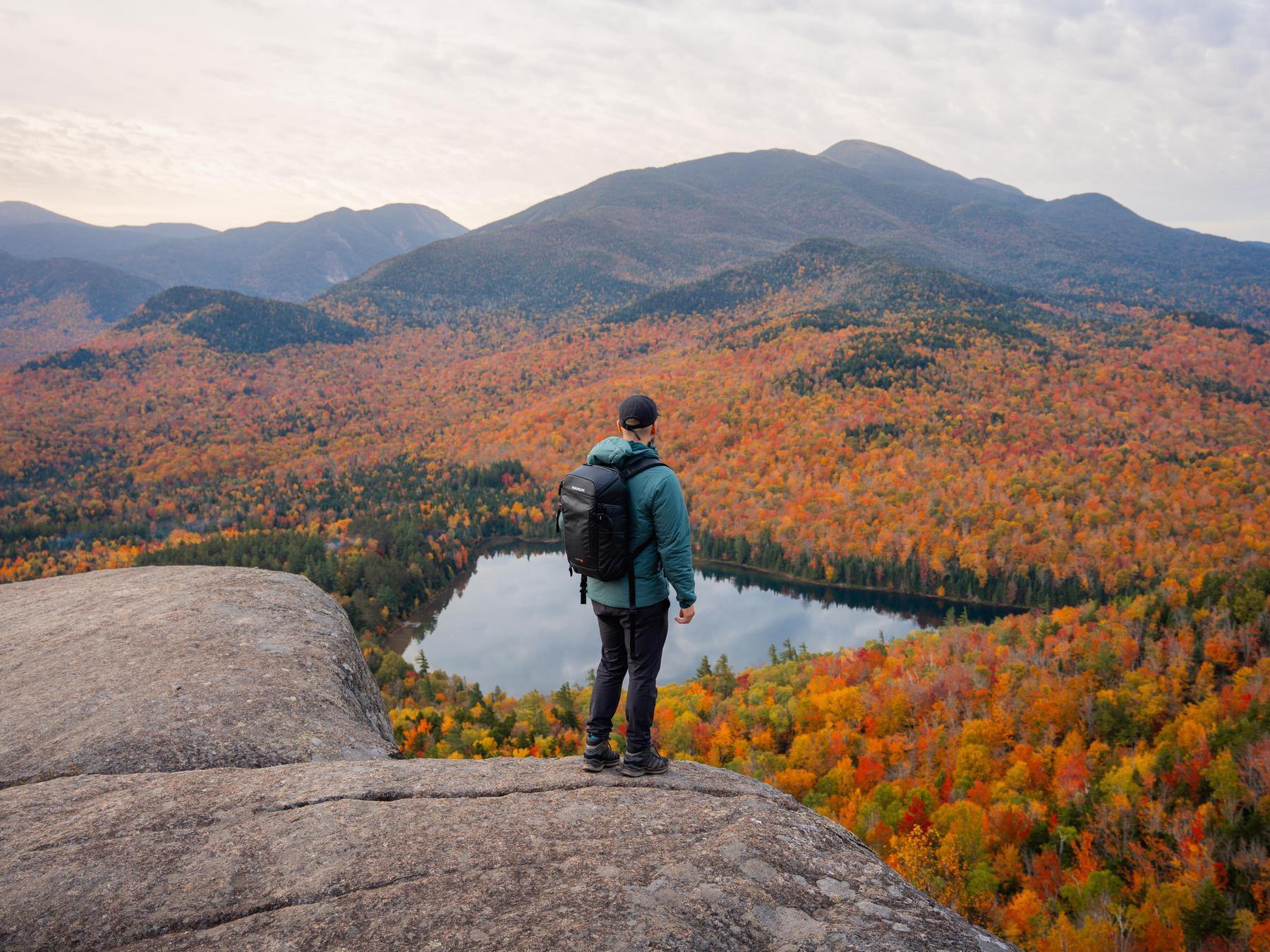 Person standing on a rocky outcrop overlooking a lake and autumn-colored forest.