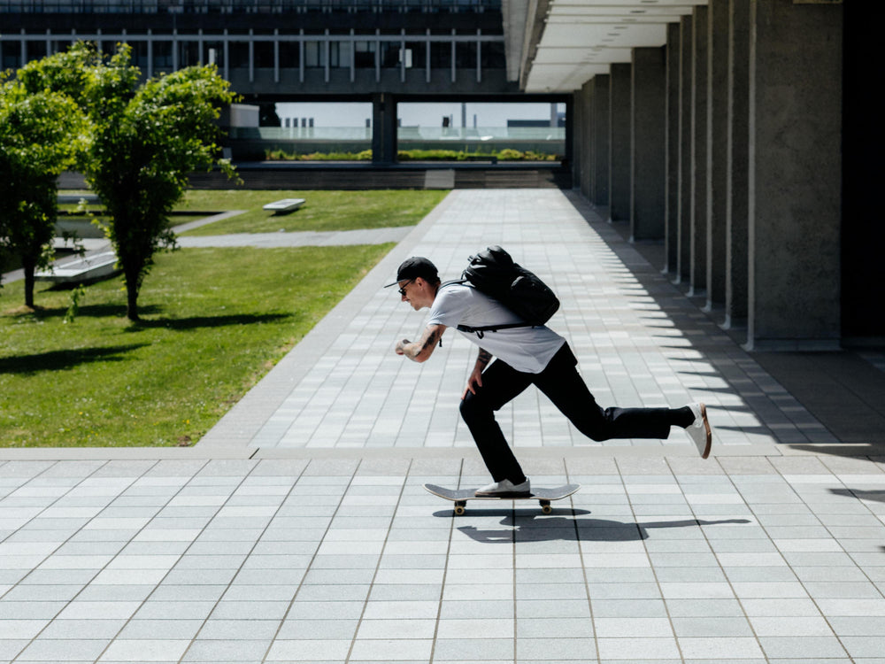 A man in a white t-shirt, black pants, and a black backpack skates on a skateboard in a modern outdoor plaza with tiled floors, grass, and columns.