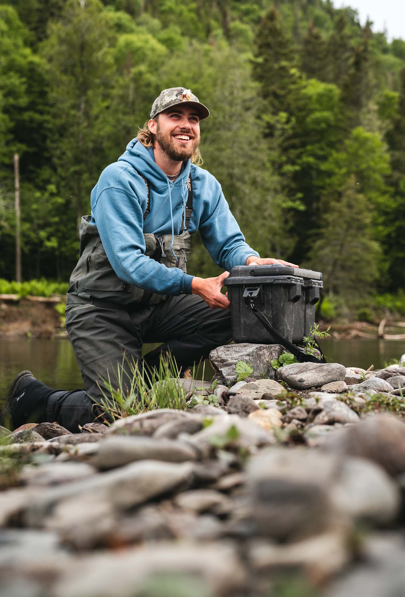 Man in waders kneeling by a river with a large black container, surrounded by greenery.