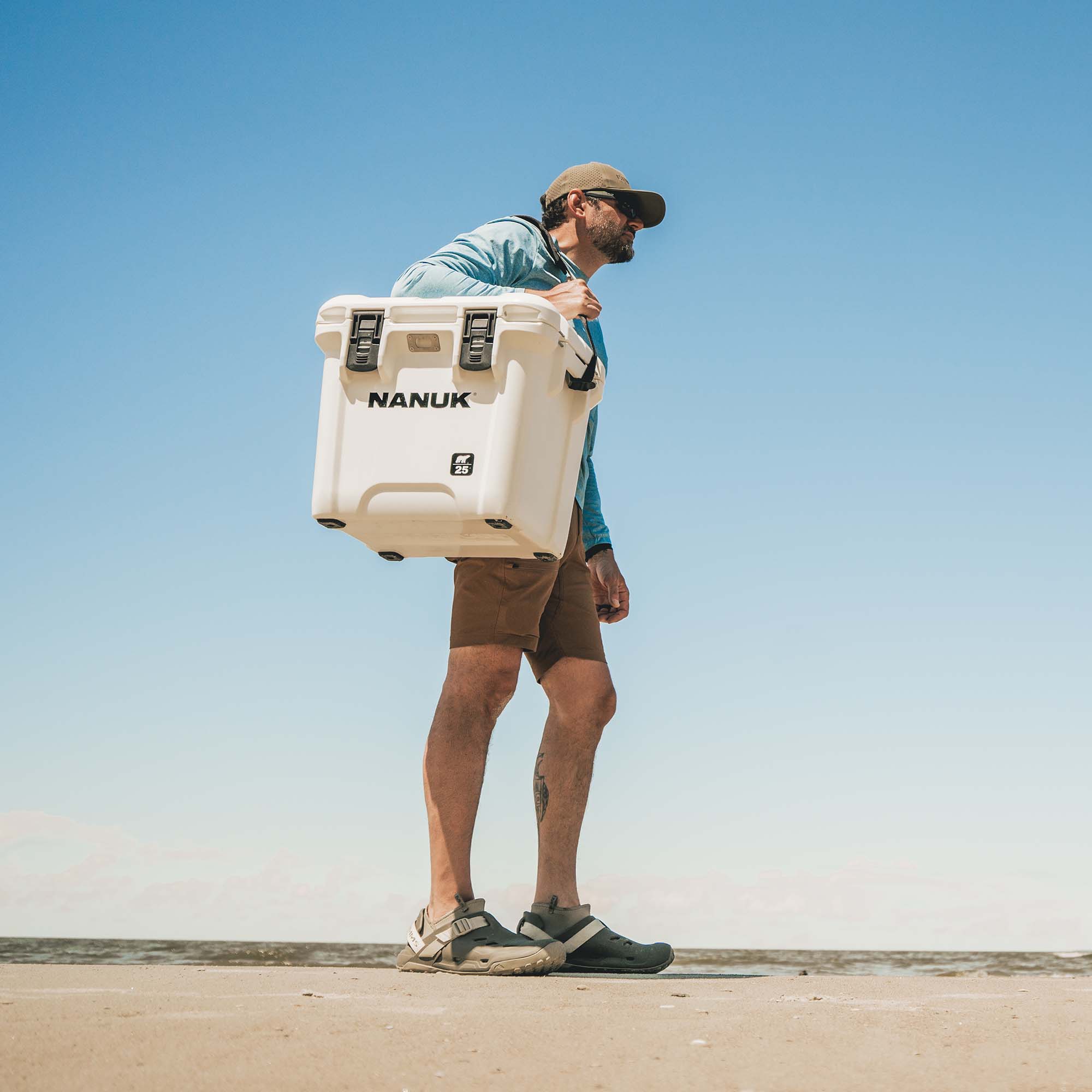 A man carrying a large white NANUK cooler on his shoulder, walking outdoors against a clear blue sky.