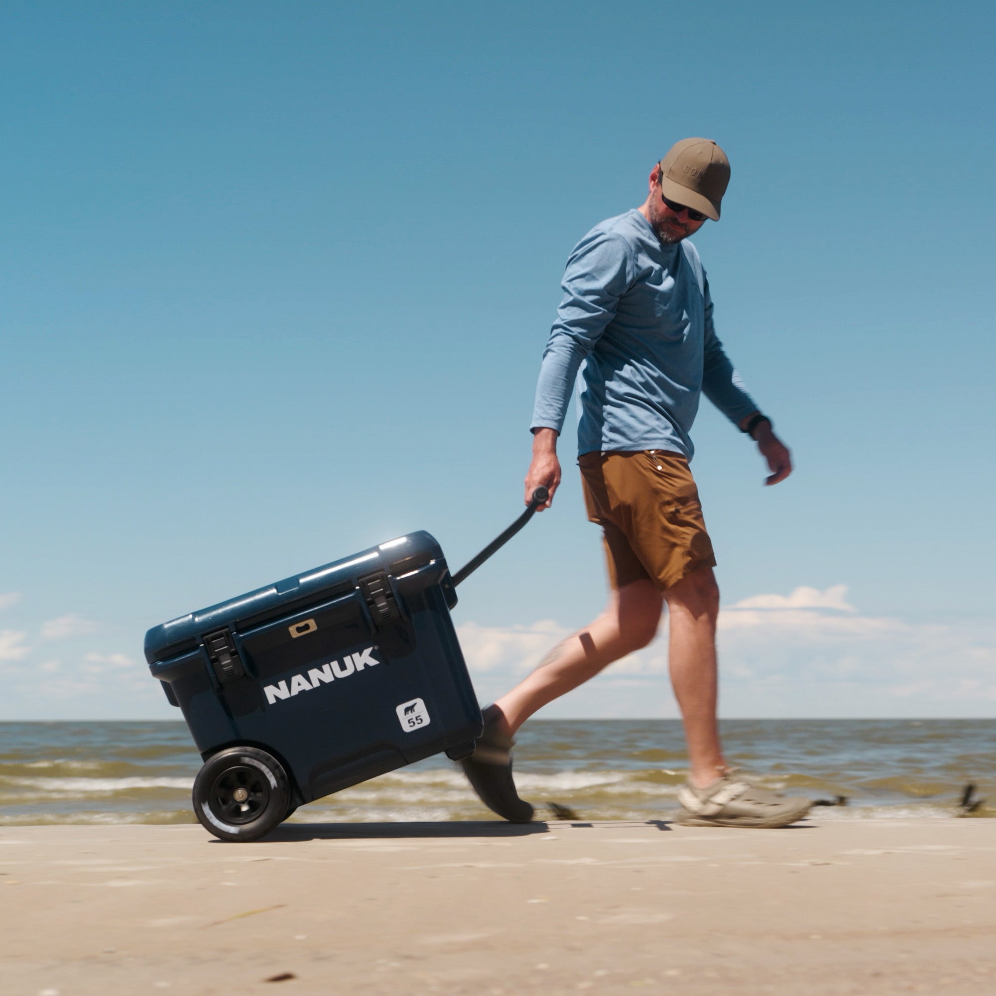 A man walks along a sandy beach pulling a dark blue Nanuk cooler on a wheeled dolly under a clear blue sky.