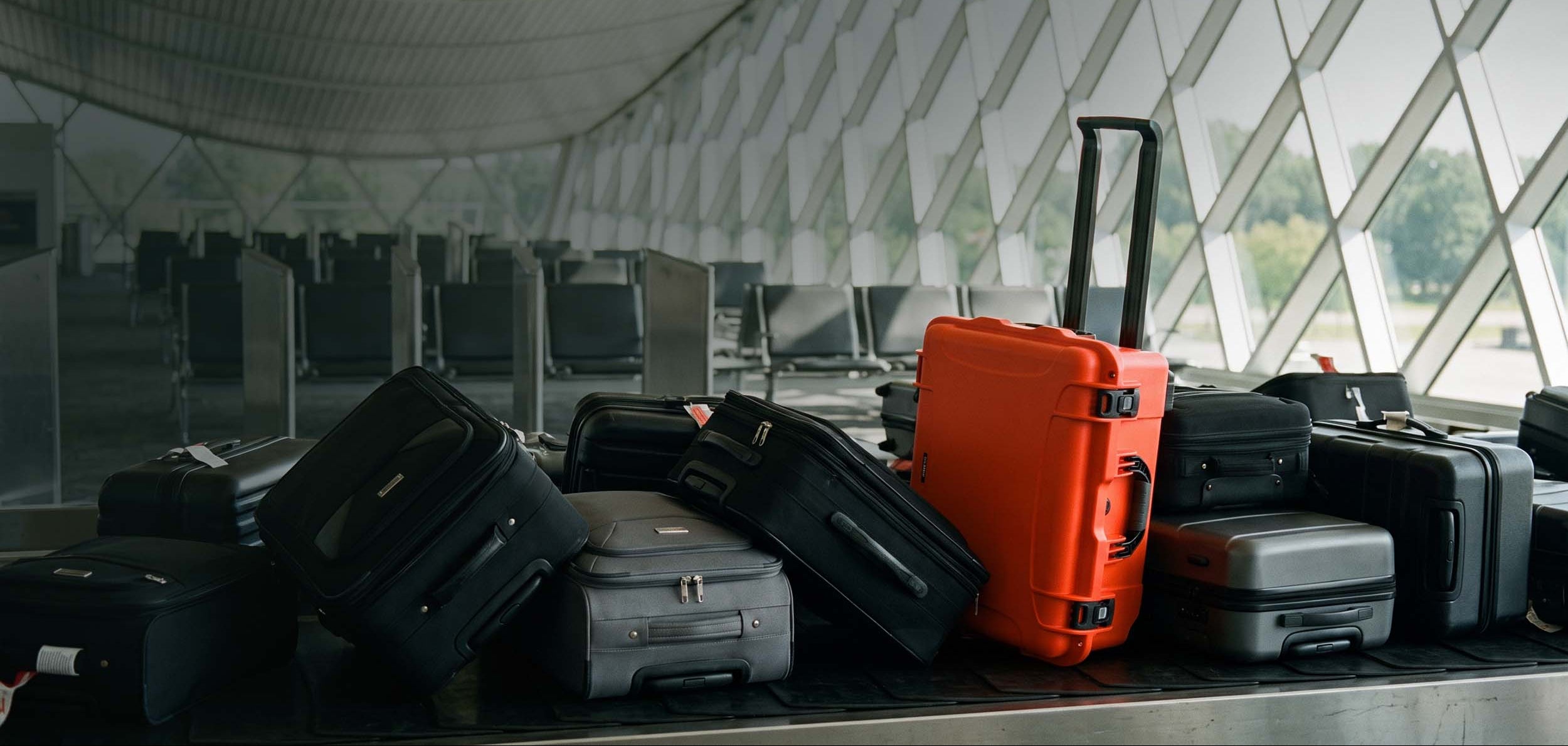 A group of hard-shell and soft-sided suitcases on a baggage carousel inside an airport terminal, with a prominent bright orange suitcase standing upright near large glass windows.