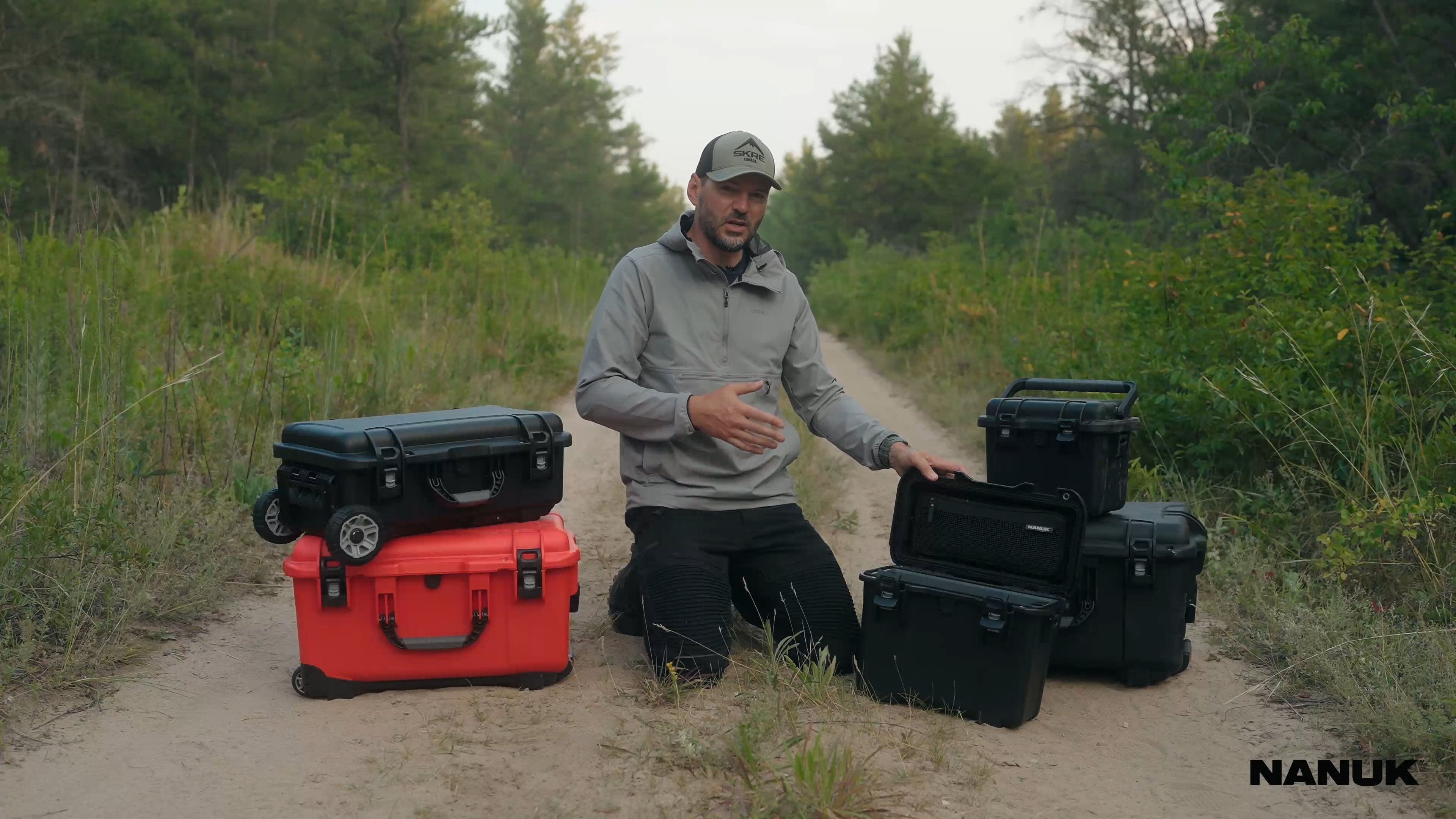 A man outdoors on a dirt path, surrounded by trees, demonstrates a collection of Nanuk protective hard cases in various sizes and colors, including black and a vibrant red. He actively showcases the cases' features, opening them to reveal organized interiors with mesh pockets, dividers, and secure compartments suitable for storing fishing lures, electronics, and other outdoor gear. The video highlights the cases' robust design, wheels for transport, and practical application, with a scene showing the man us